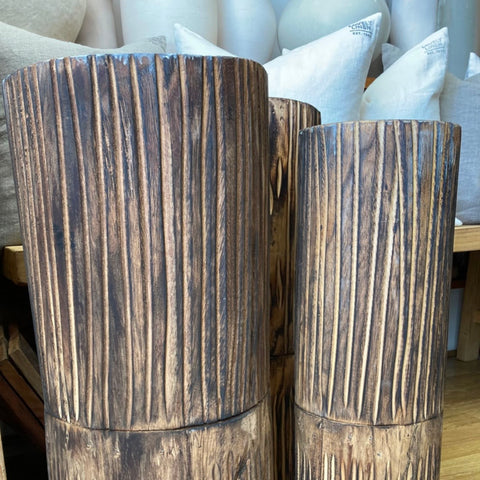 Two rustic dark brown wooden vases with a vertically grooved, textured finish, shown in a home decor setting with neutral-toned pillows in the background.